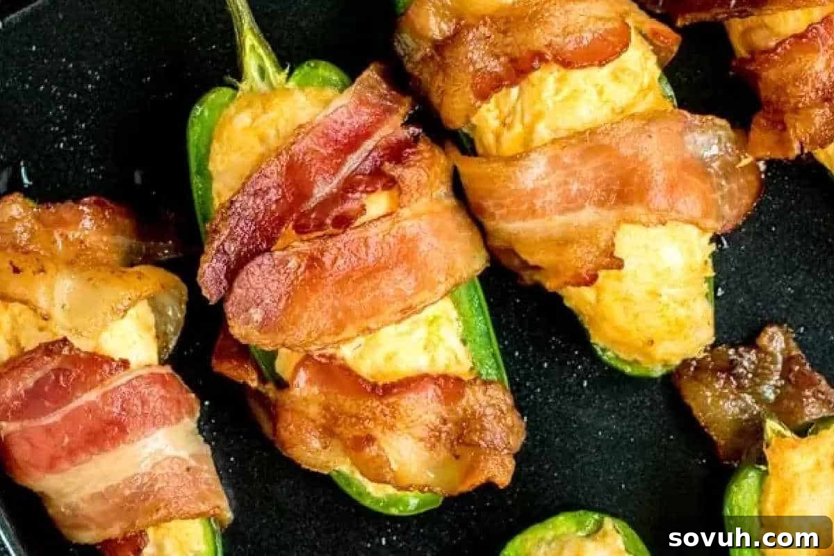Close-up of vibrant jalapeño halves, generously stuffed with a creamy cheese filling infused with buffalo chicken, and expertly wrapped in crispy, golden-brown bacon, presented on a dark surface.