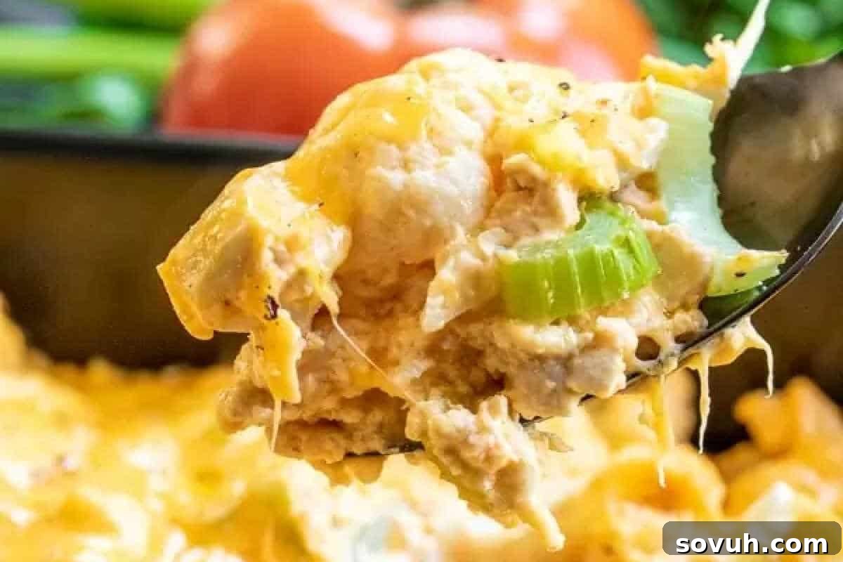 A close-up of a generous spoonful of bubbling, cheesy buffalo chicken casserole, rich with pieces of tender chicken, cauliflower florets, and melted cheese, with fresh vegetables blurred in the background.