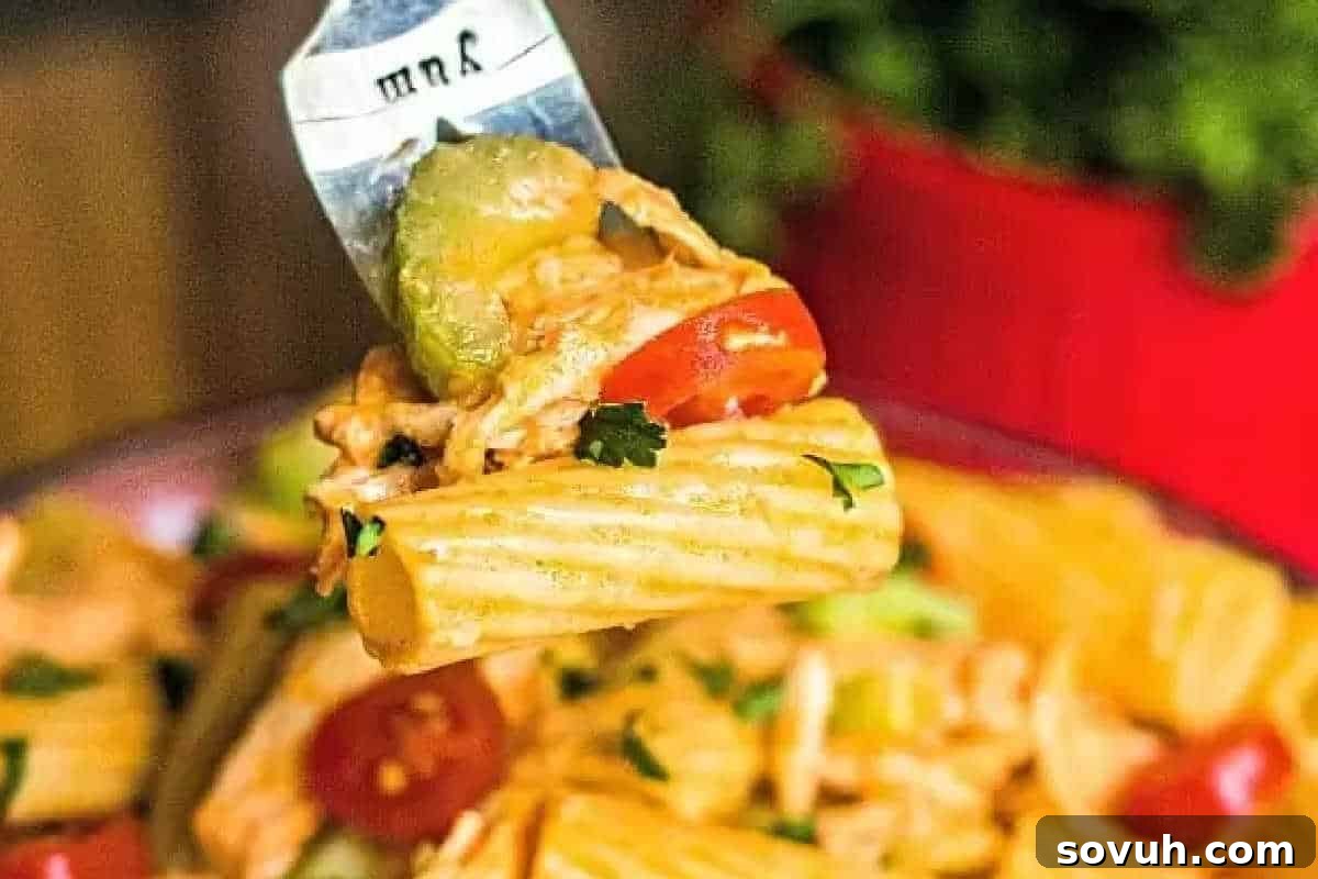 A fork lifts rigatoni pasta, beautifully coated with buffalo sauce, mixed with tender shredded chicken, bright cherry tomatoes, crisp zucchini, and fresh herbs, with more vibrant pasta salad visible in the background.