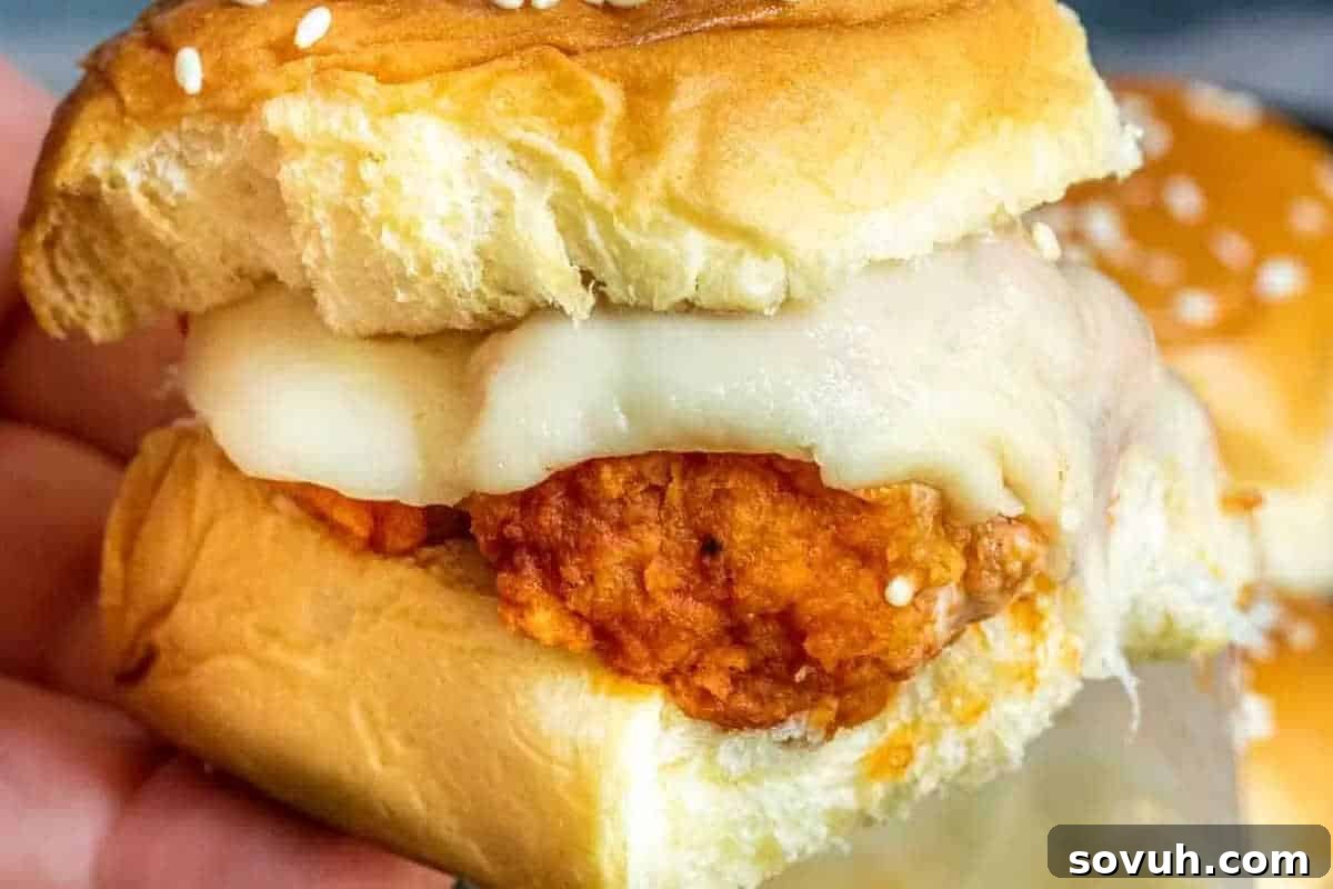 A hand holds a perfectly constructed slider sandwich, featuring a golden-brown breaded chicken patty smothered in buffalo sauce, topped with melted provolone cheese, all nestled in a soft, sesame seed bun.