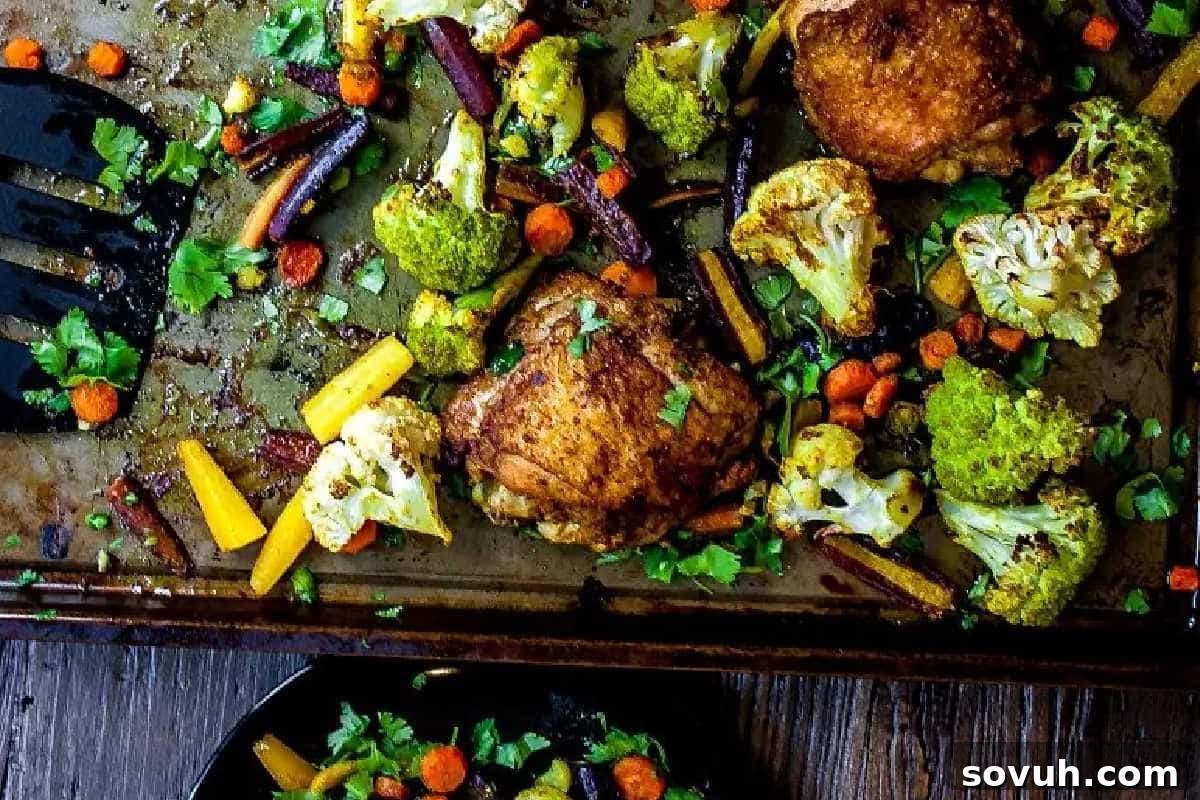 Roasted chicken thighs with carrots, broccoli, and cauliflower on a baking sheet, garnished with fresh herbs. Some food is served in a black dish at the bottom.