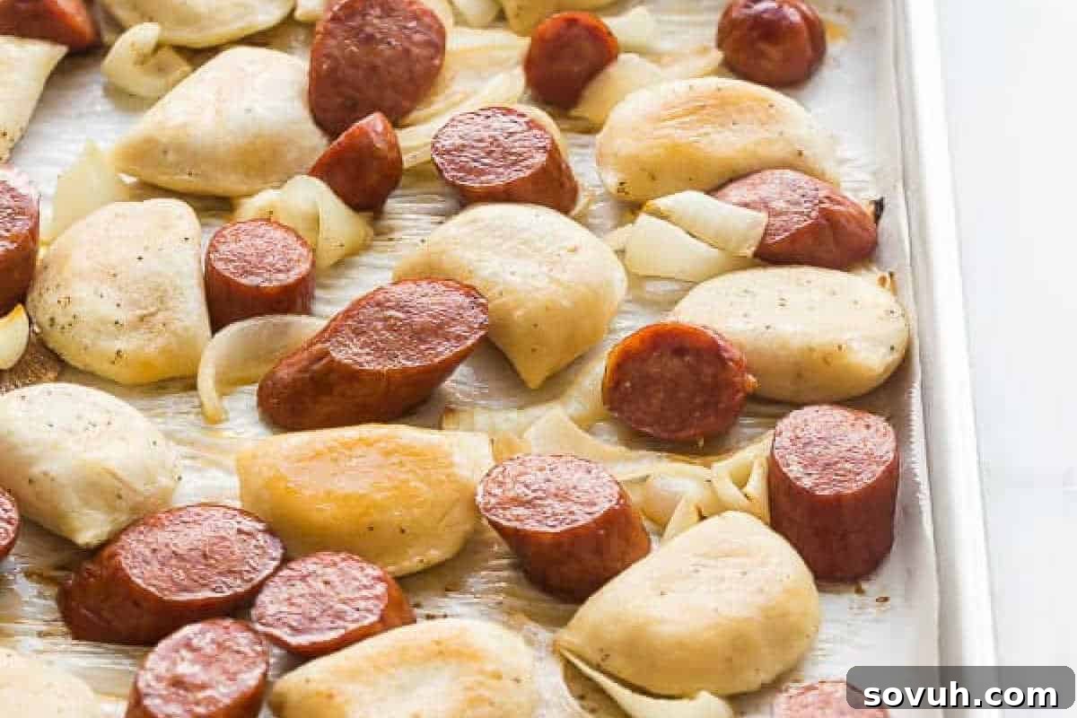 Sliced sausage, pierogies, and chopped onions arranged on a metal baking sheet, ready for roasting.