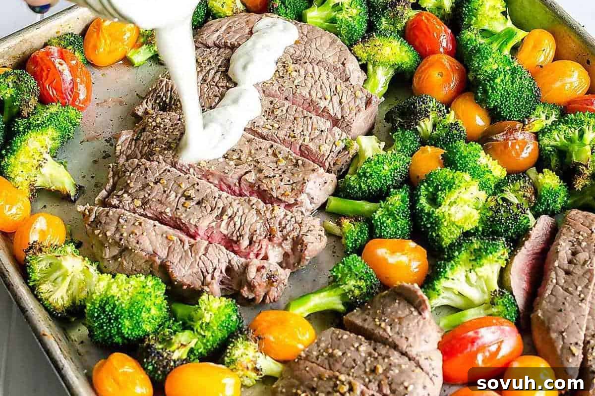 Sliced steak on a sheet pan surrounded by broccoli and cherry tomatoes, with a creamy white sauce being poured over the steak.
