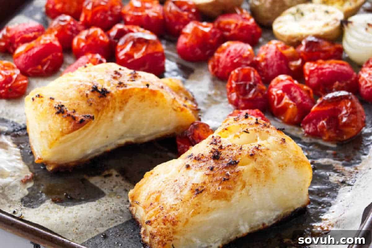 Two pieces of baked white fish and roasted cherry tomatoes on a metal baking tray, with some roasted potato slices and onion visible in the background.