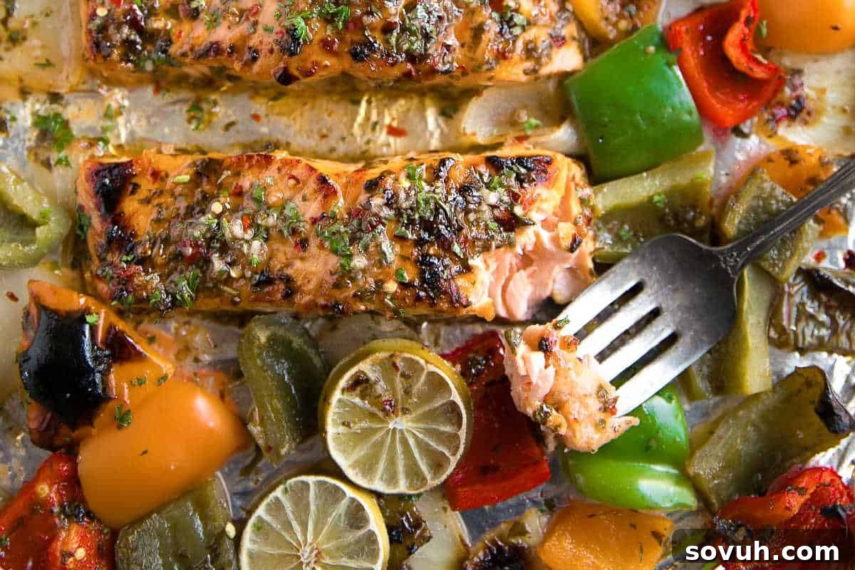 Baked salmon fillets topped with herbs, surrounded by roasted bell peppers and lime slices, with a fork holding a bite of salmon.