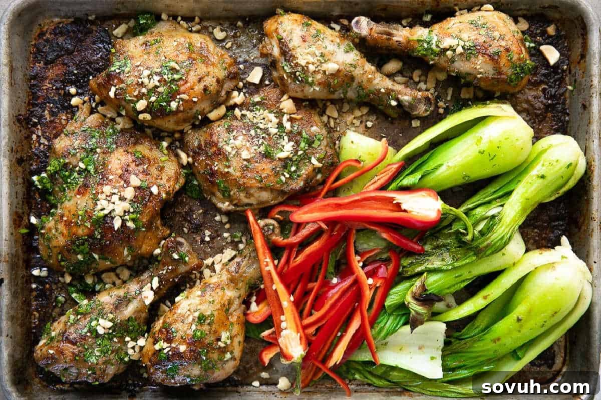 Baked chicken drumsticks and thighs garnished with herbs and nuts, served with sliced red bell peppers and baby bok choy on a baking sheet.