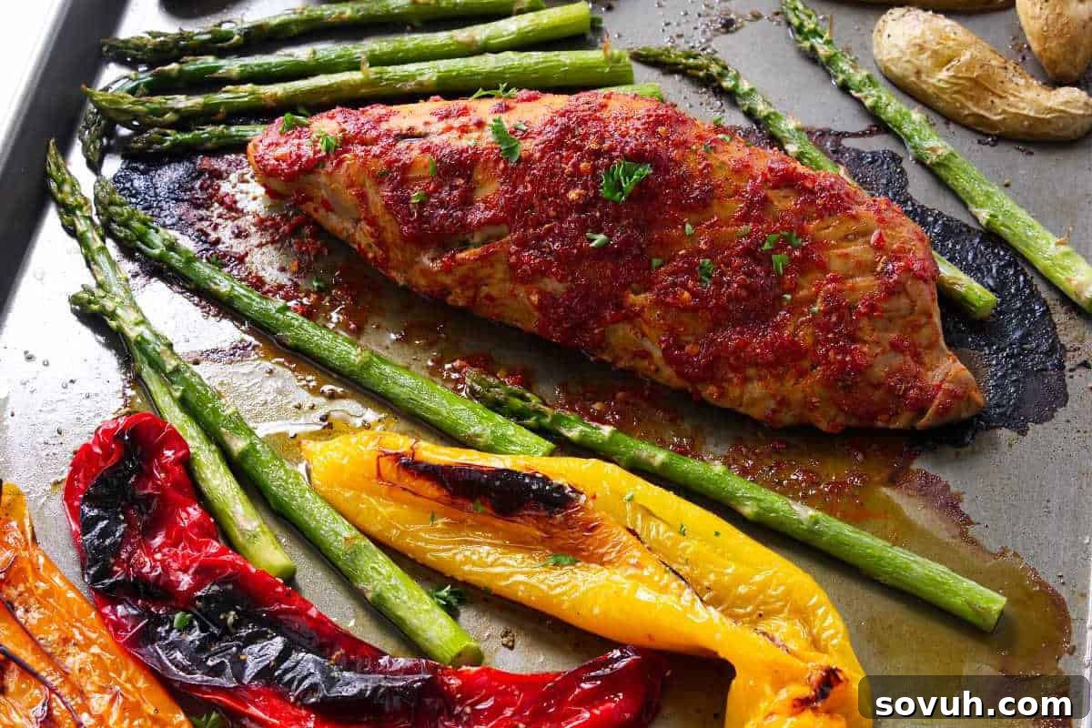 A seasoned baked chicken breast on a sheet pan, surrounded by roasted asparagus, red and yellow bell peppers, and potato halves.