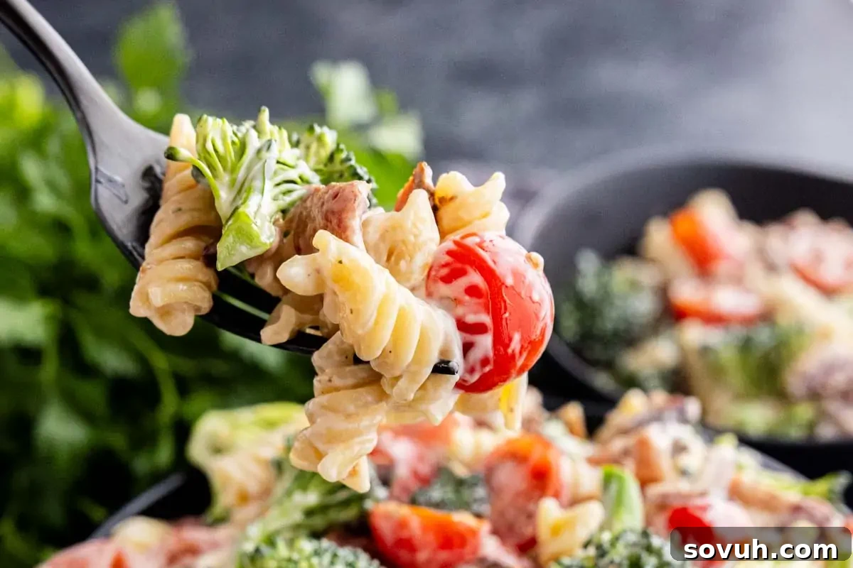 A close-up of a fork holding creamy rotini pasta with broccoli, cherry tomatoes, and pieces of crispy bacon, with a large bowl of the salad blurred in the background, hinting at a delicious, savory dish.