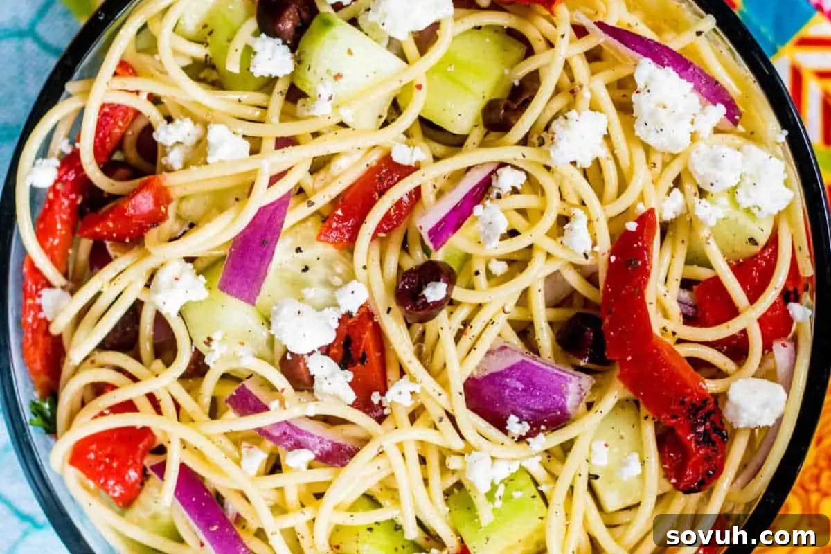 A fresh Greek Spaghetti Salad in a rustic bowl, featuring long spaghetti strands, crumbled feta cheese, kalamata olives, roasted red peppers, and sliced cucumbers, all dressed in a vibrant Greek vinaigrette.