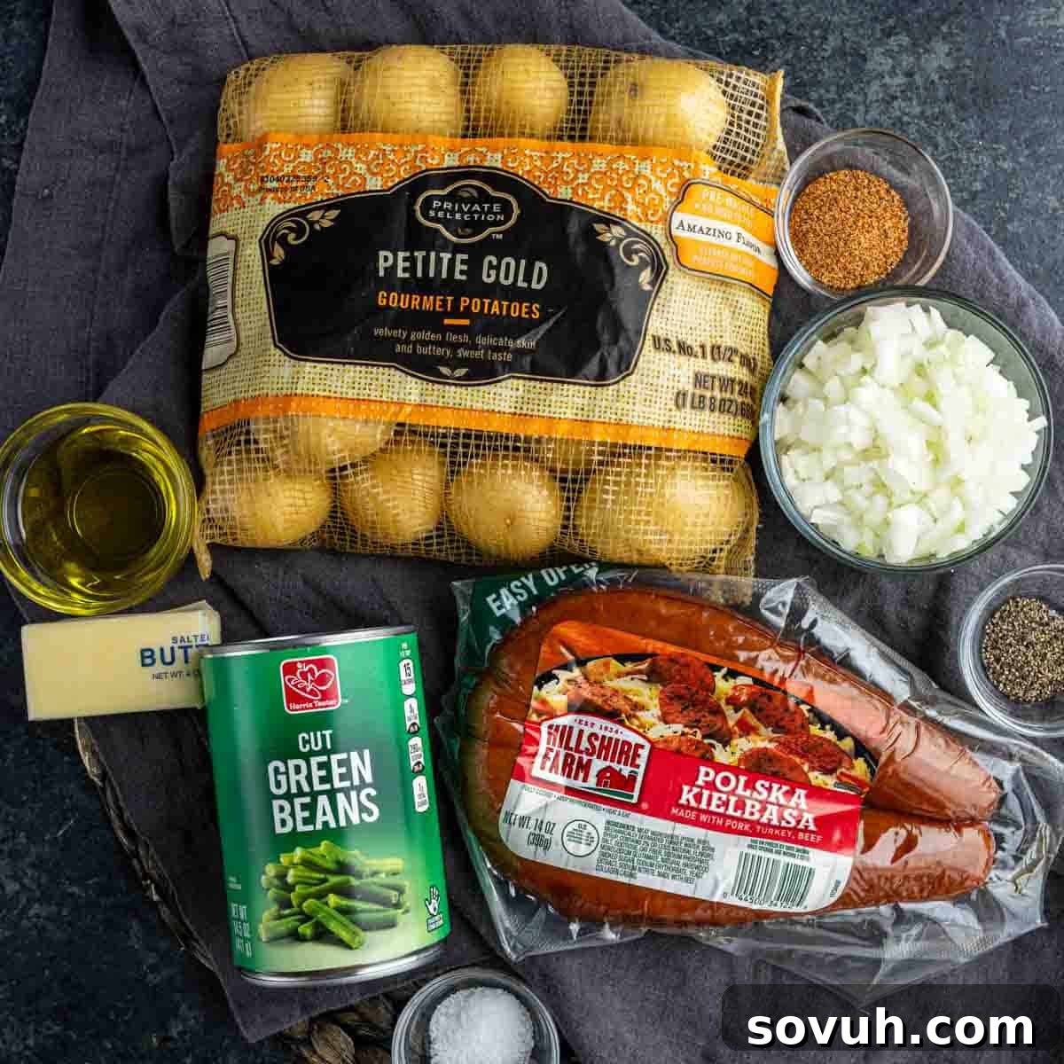 Hearty Sausage, Potato, and Green Bean Bake 3 Top-down view of ingredients: petite gold potatoes, green beans, Polska kielbasa sausage, diced onions, butter, olive oil, and assorted seasonings on a dark surface.