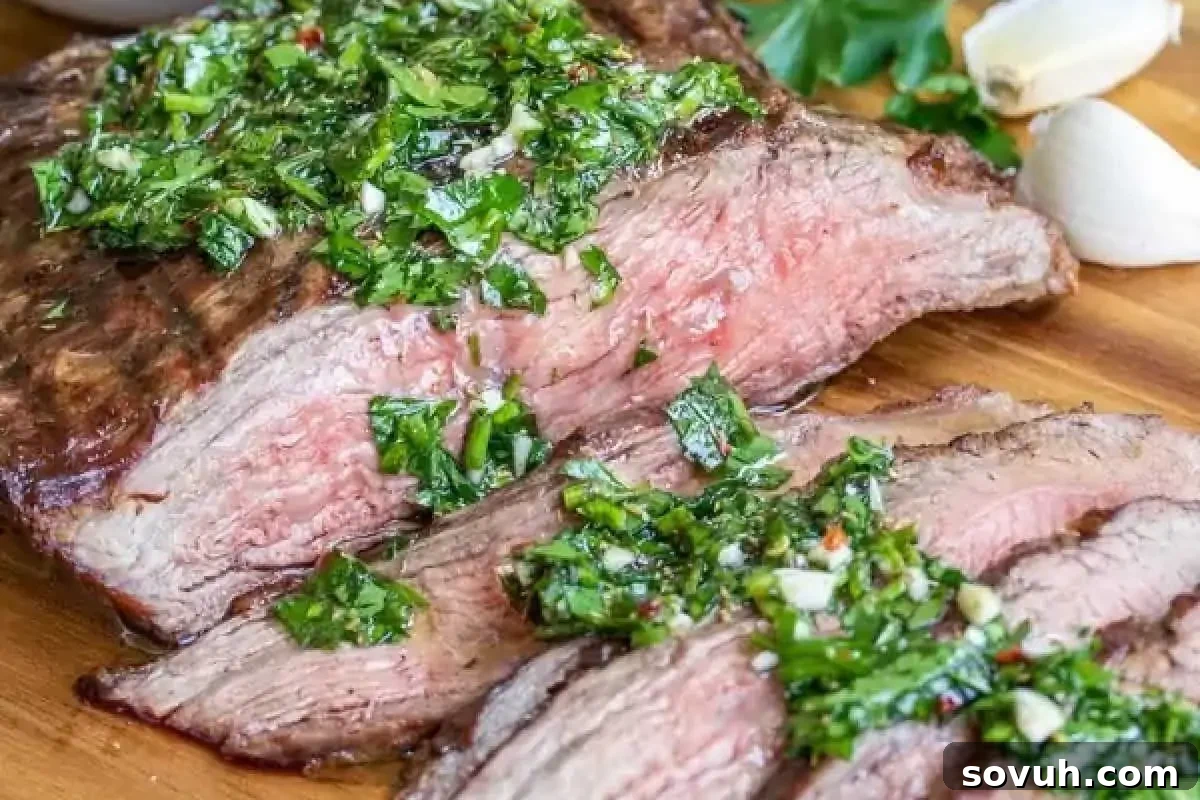 Sliced medium-rare steak topped with green herb salsa on a wooden cutting board, accompanied by garlic cloves and fresh herbs.