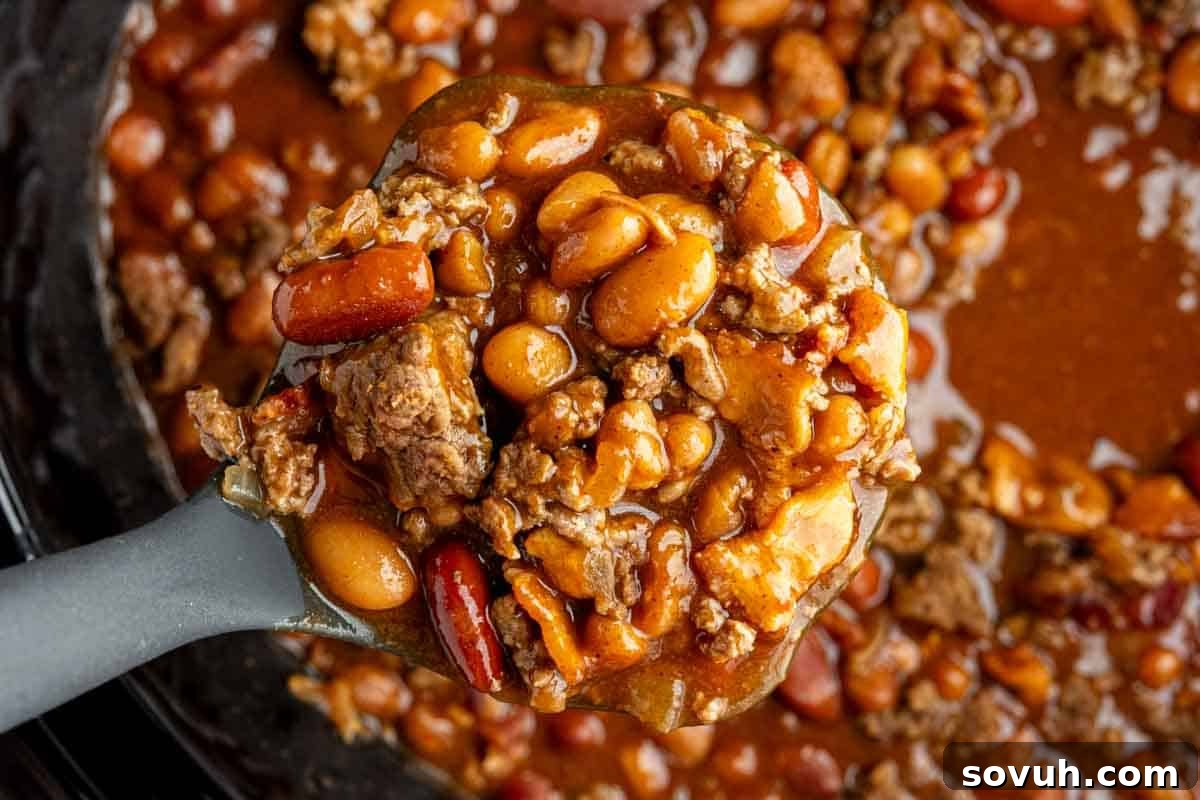 A close-up of a ladle holding a mixture of crock pot cowboy baked beans—baked beans, ground beef, and sauce—over a pot of the same hearty cowboy beans, showcasing its rich texture and ingredients.