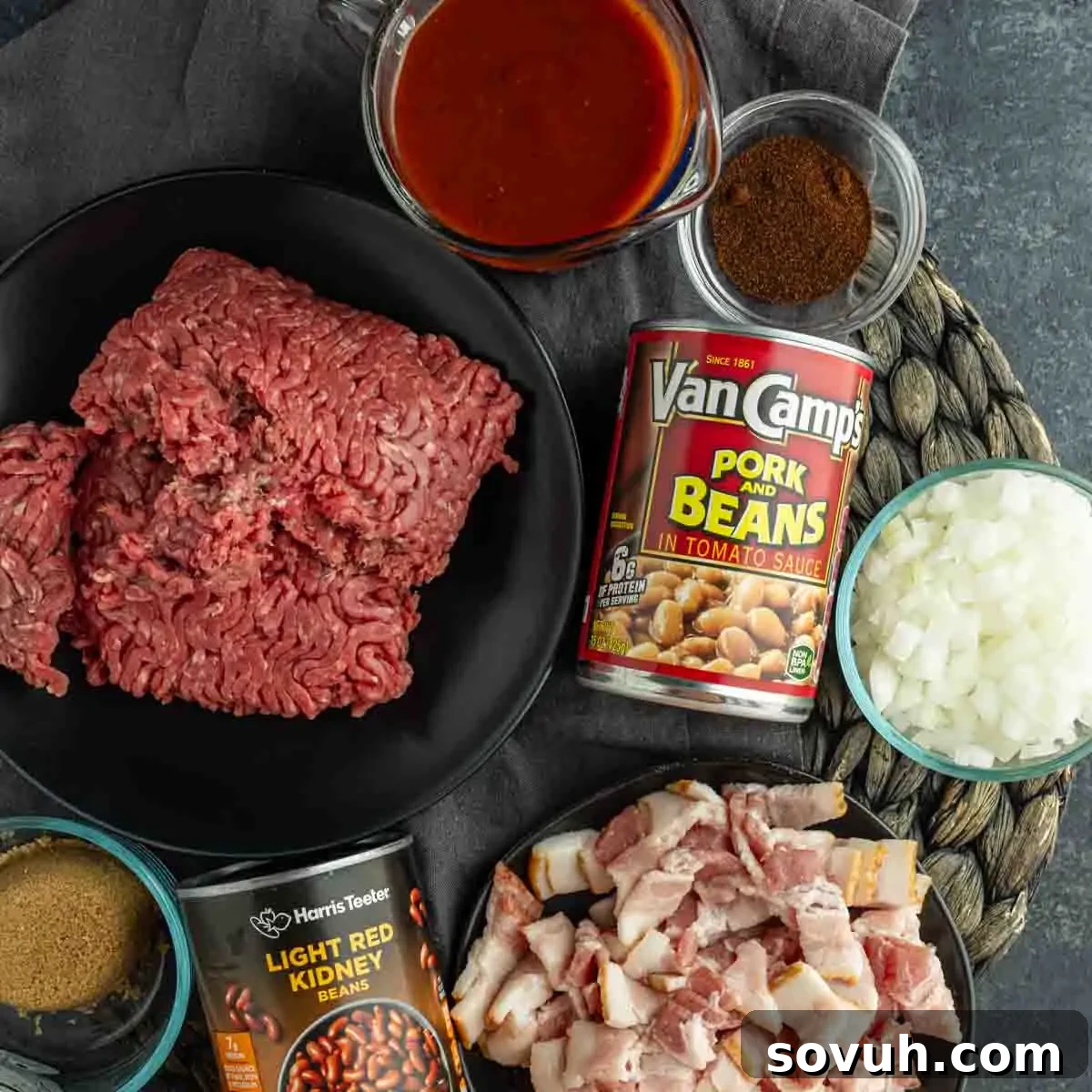 A flat lay of fresh ingredients for crock pot cowboy baked beans, including lean ground beef, canned pork and beans, kidney beans, diced onions, crispy bacon pieces, chili powder, rich brown sugar, and tangy tomato sauce, ready for slow cooking.