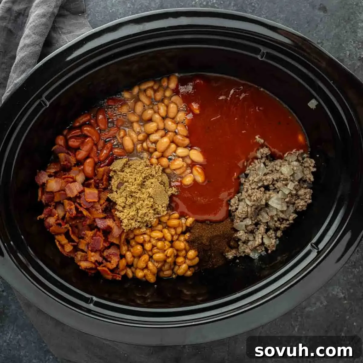 A slow cooker filled with uncooked ingredients for cowboy beans, including ground beef, crispy bacon, various beans, barbecue sauce, brown sugar, and seasonings, arranged separately before mixing for a convenient crock pot recipe.