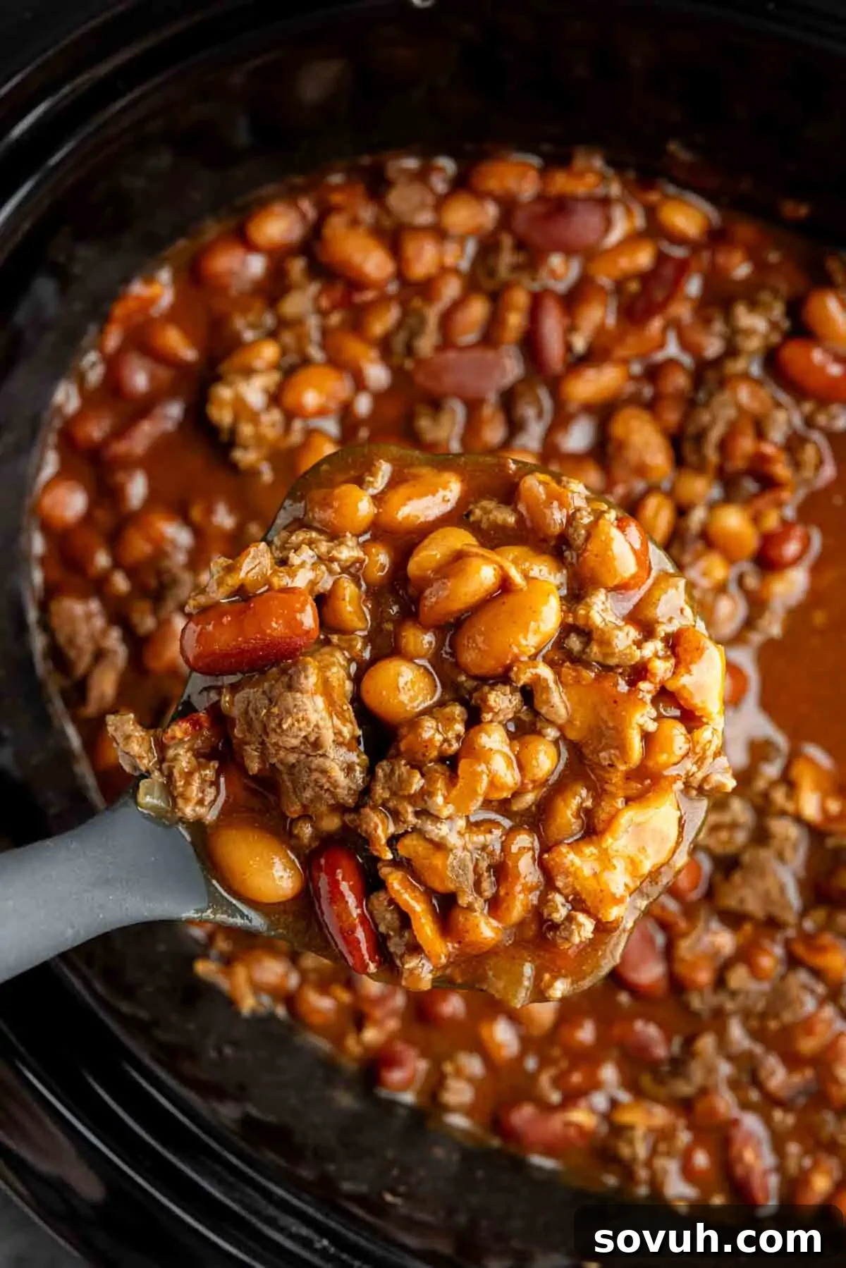 A close-up of a ladle scooping hearty cowboy beans with ground beef and a medley of mixed beans from a crockpot, illustrating the rich, saucy texture of this easy crock pot recipe.