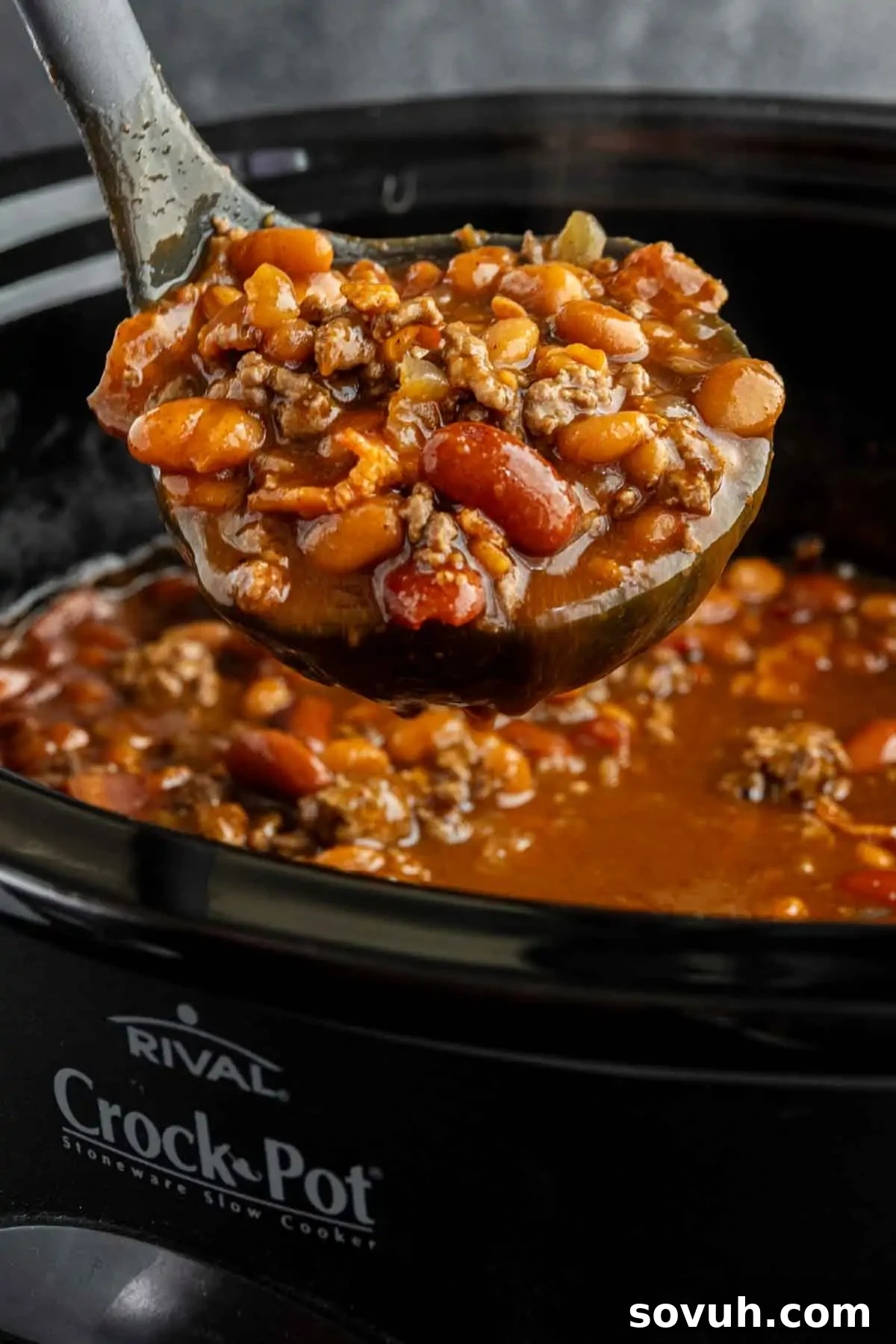 A ladle scoops hearty cowboy beans with ground meat and savory spices from a Crock-Pot slow cooker, highlighting the rich, thick sauce.