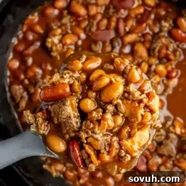 A close-up of a spoonful of chili with ground beef, kidney beans, and pinto beans over a pot of chili, illustrating the texture of the cowboy beans.