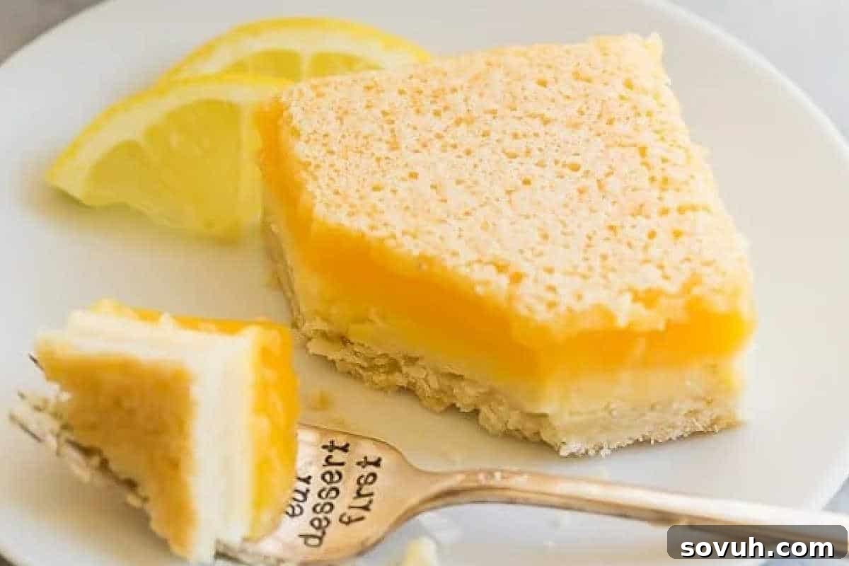 A square slice of lemon bar with a layered crust and yellow filling sits on a white plate with two lemon slices; a fork holds a small piece of the dessert.