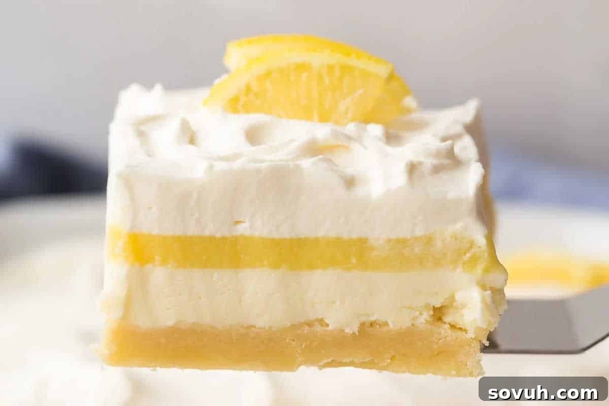 A close-up of a layered lemon dessert bar with a shortbread crust, creamy filling, lemon layer, whipped topping, and a lemon slice garnish.
