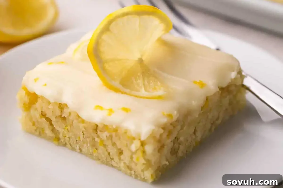 A square slice of lemon cake with white icing, garnished with a lemon slice, on a white plate with a fork.