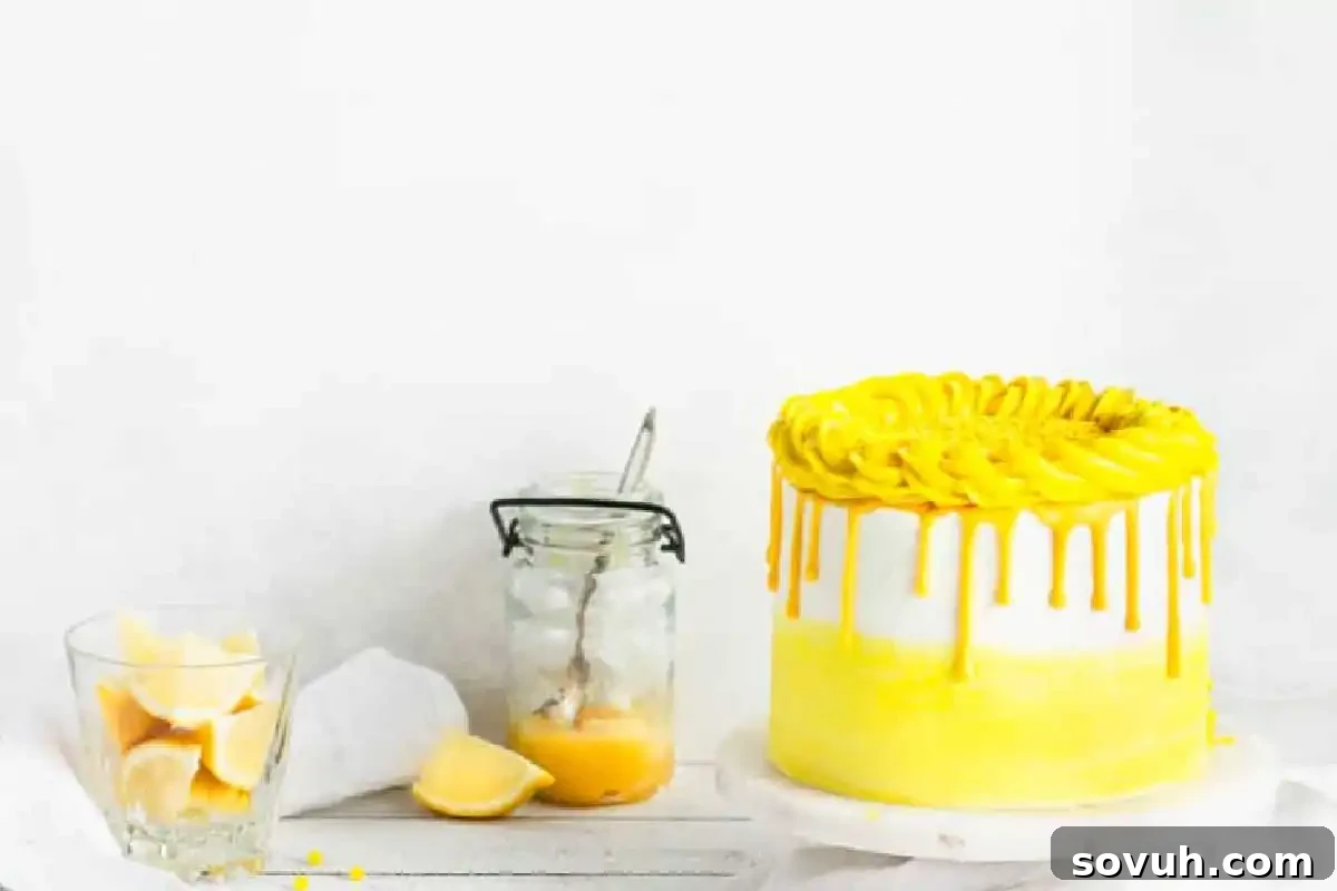 A yellow and white frosted cake with yellow drizzle sits on a stand next to a jar of lemon curd, a spoon, lemon wedges in a glass, and a lemon slice on a white surface.