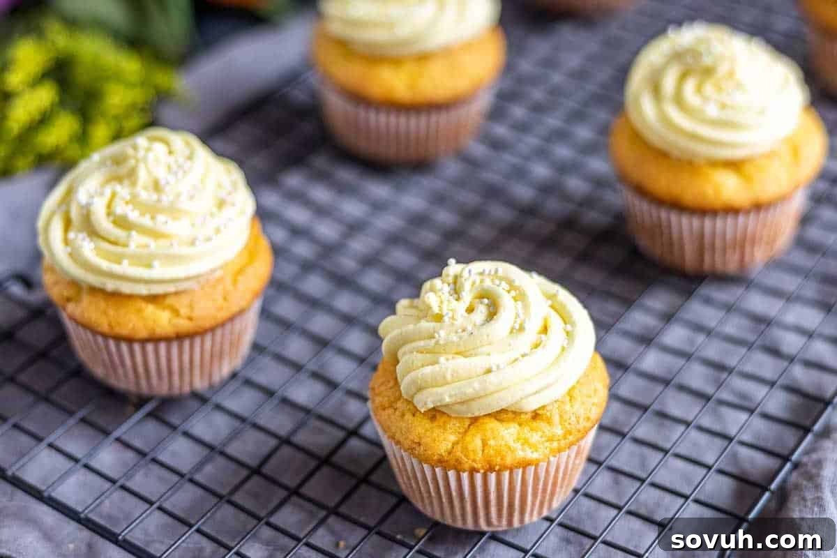 Four vanilla cupcakes with swirled frosting and white sprinkles sit on a cooling rack.