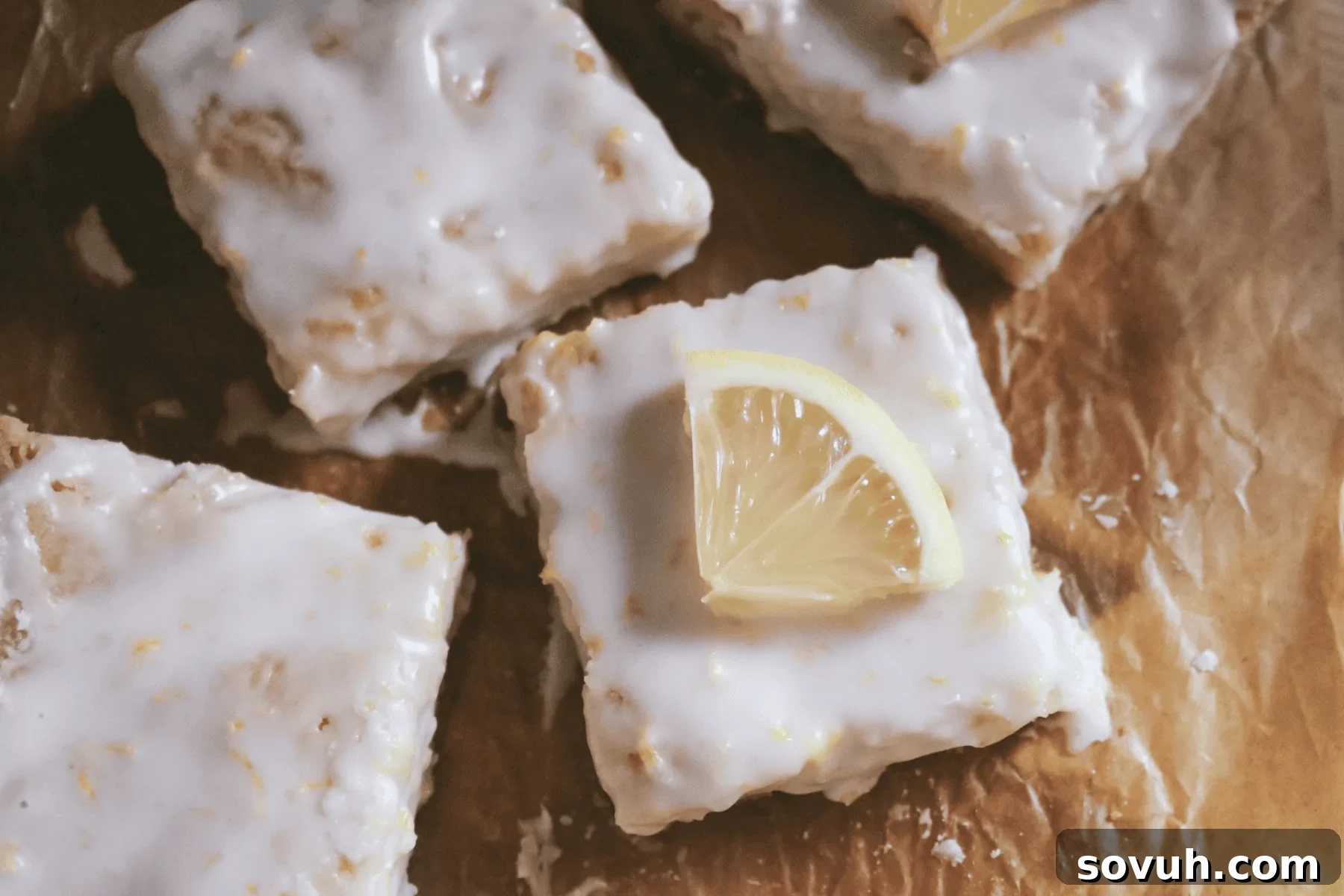 Four lemon bars with white icing are arranged on brown parchment paper; one bar is topped with a lemon slice.