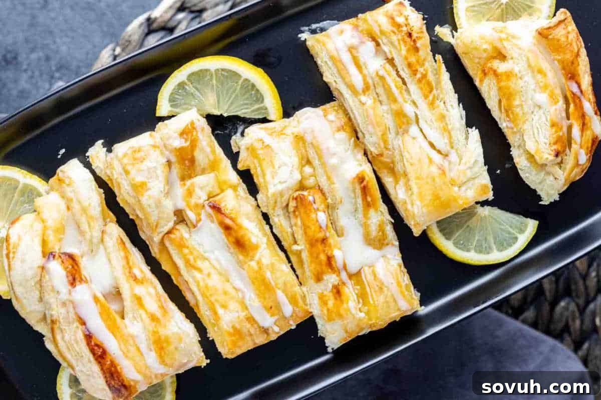 Five rectangular pieces of glazed pastry are arranged on a black plate with lemon slices placed around them.