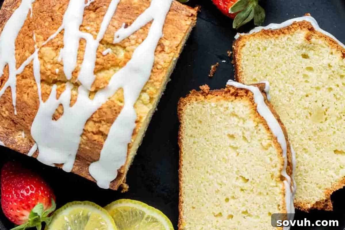 A loaf of glazed lemon bread on a black surface, with two slices cut and placed beside it. Fresh strawberries and lemon slices are arranged nearby.