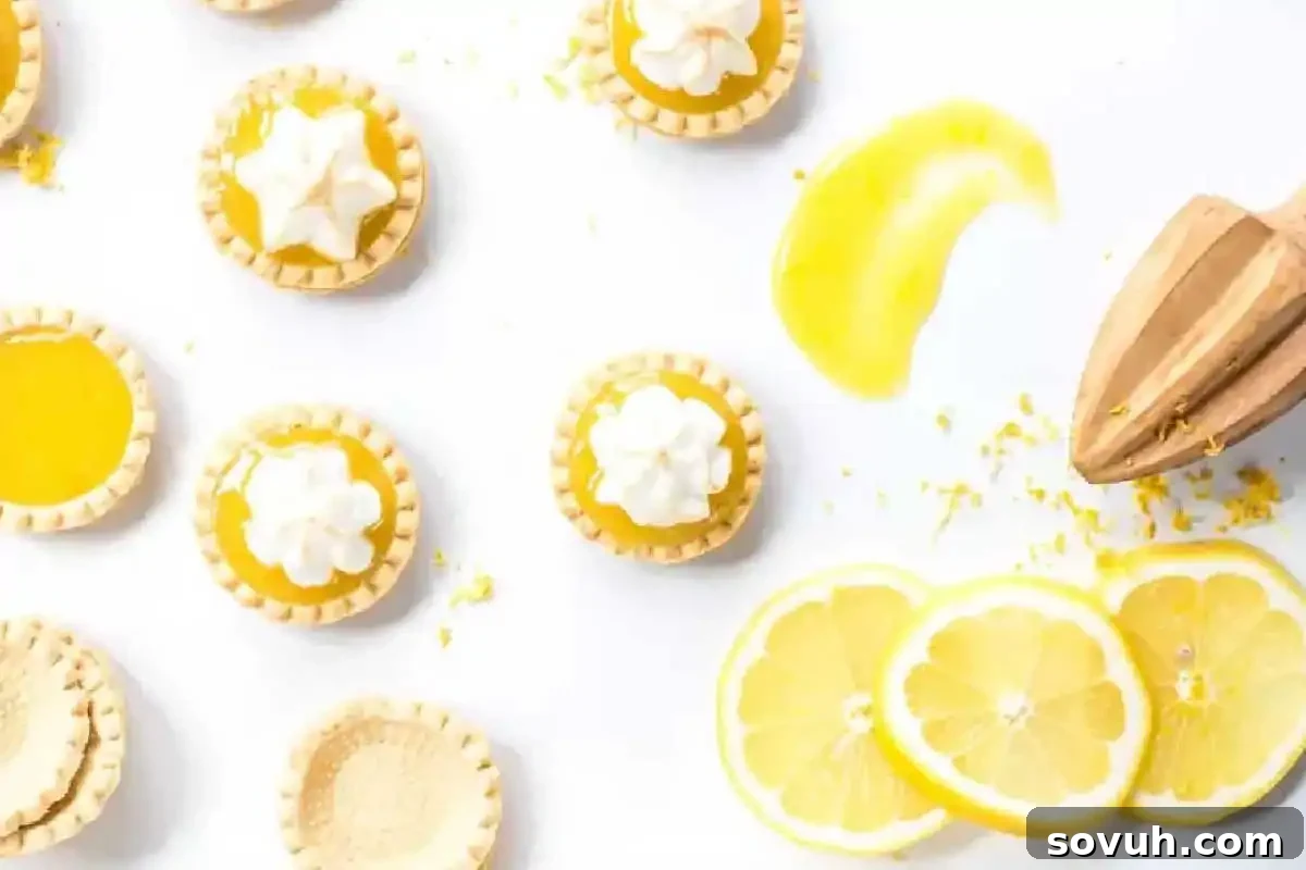 Mini lemon tarts and tart shells are arranged on a white surface with lemon slices, lemon zest, a wooden citrus reamer, and a splash of lemon curd.