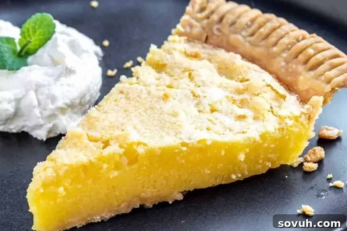 A slice of yellow custard pie with a crumbly crust is served on a black plate next to a dollop of whipped cream garnished with a mint leaf.