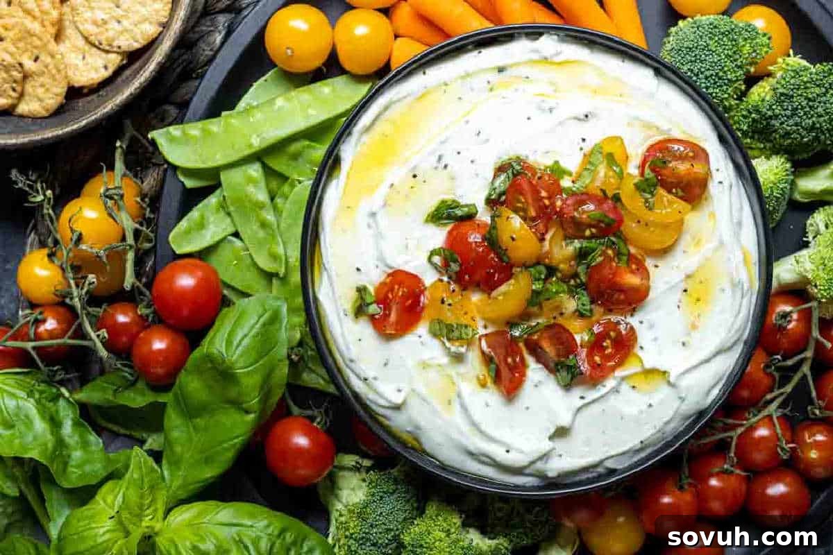 A bowl of creamy dip topped with chopped tomatoes and herbs, surrounded by fresh vegetables including snap peas, cherry tomatoes, carrots, and broccoli.