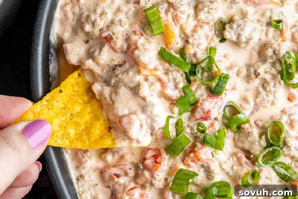 A hand dips a tortilla chip into a creamy cheese dip with ground meat, diced tomatoes, and sliced green onions.