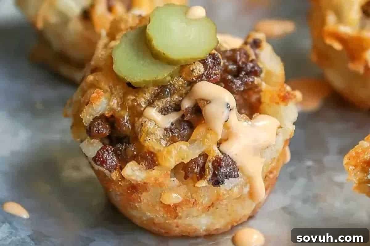 Close-up of a savory muffin topped with ground meat, melted cheese, sliced pickles, and a drizzle of sauce on a metal tray.