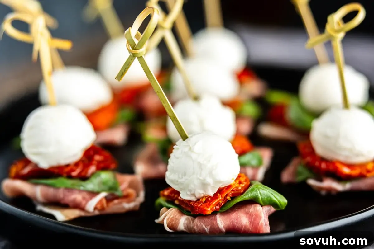Skewered appetizers with mozzarella balls, sun-dried tomatoes, basil leaves, and prosciutto on a black plate.