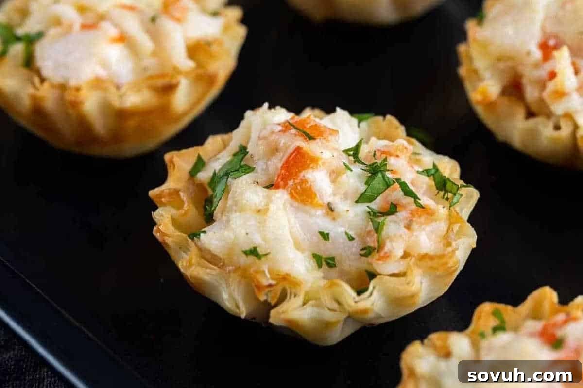 Close-up of mini phyllo pastry cups filled with creamy crab mixture, garnished with chopped herbs, on a dark surface.