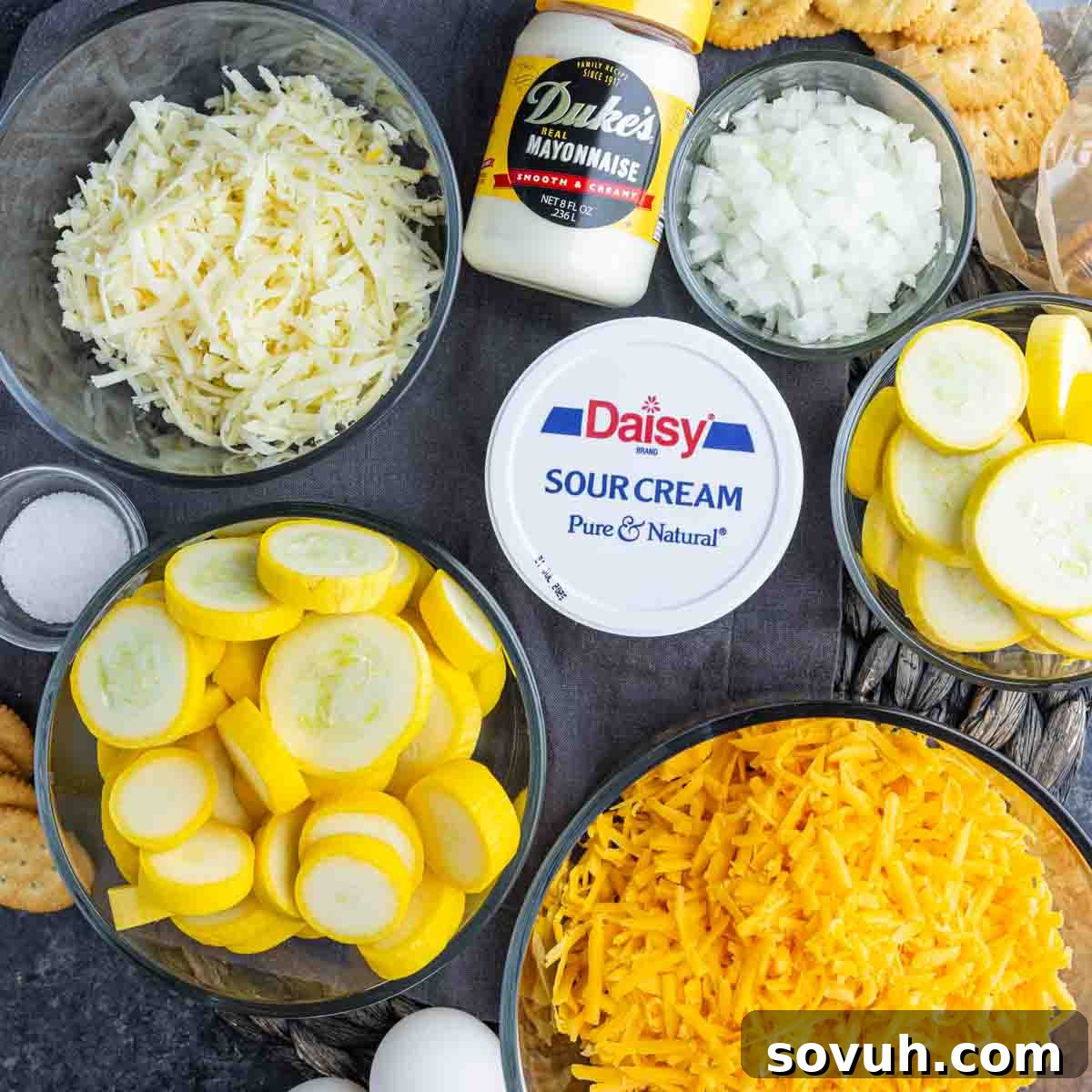 Bowls arrayed on a countertop, holding the fresh ingredients for a classic southern yellow squash casserole, including shredded cheddar and Swiss cheese, chopped yellow onions, thinly sliced yellow squash, creamy mayonnaise, sour cream, salt, a whisked egg, and crushed Ritz crackers.