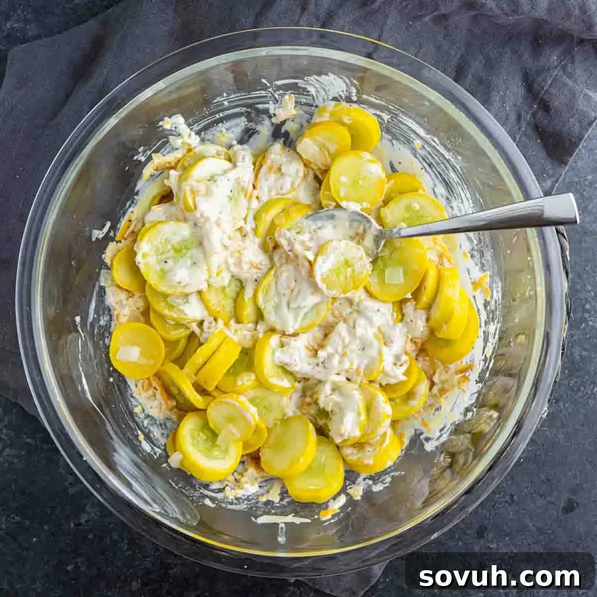 A glass mixing bowl filled with a creamy yellow squash mixture, ready for baking into an easy yellow squash casserole. A metal spoon is resting inside the bowl, which is placed on a dark surface.