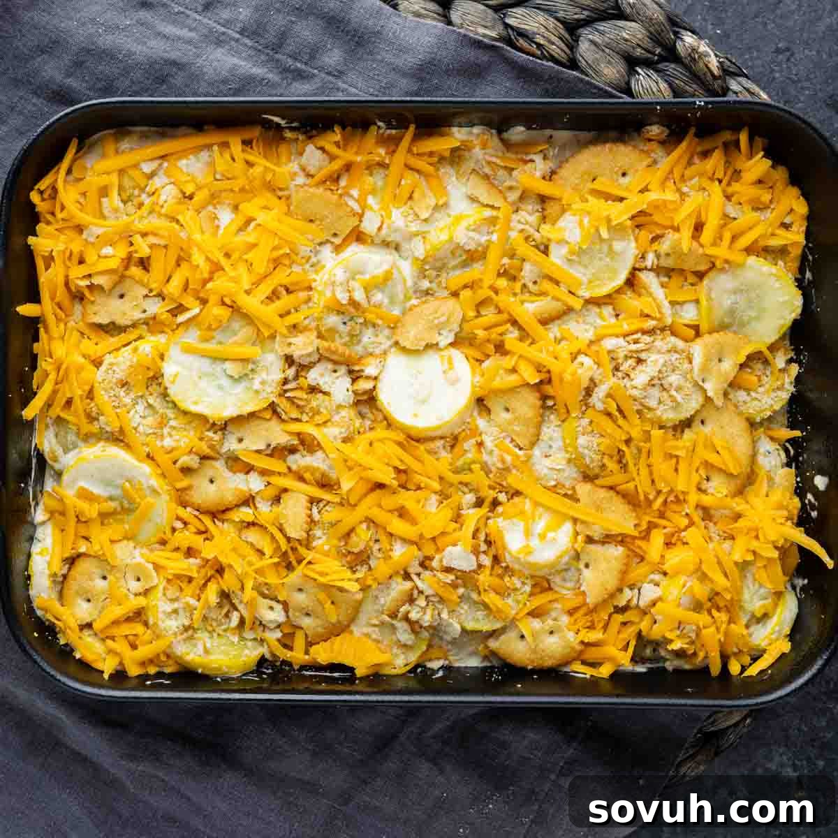 A rectangular baking dish is filled with layers of sliced yellow squash, creamy cheddar cheese sauce, and crumbled crackers, forming the base for an easy yellow squash casserole, set on a dark patterned cloth.
