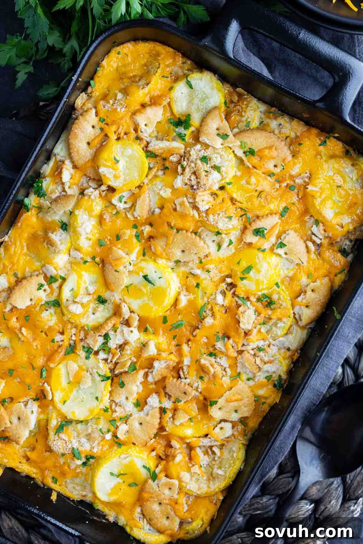 A beautifully baked classic Southern yellow squash casserole, featuring a melted cheese topping, crushed Ritz crackers, and a sprinkle of fresh parsley, served in a rectangular black baking dish on a rustic surface.