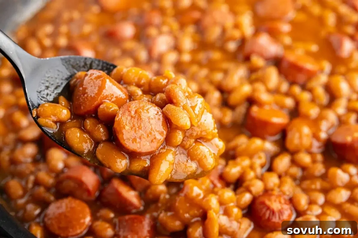 A tantalizing close-up of a spoonful of classic baked beans and perfectly sliced sausage nestled in a rich, thick sauce, with more beans and sausage invitingly visible in the background.