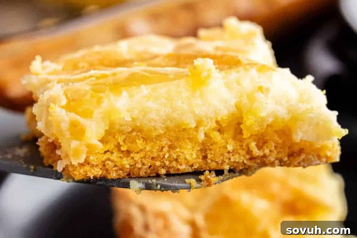 A close-up of a square slice of gooey butter cake with a golden crust and soft, creamy topping on a spatula, promising rich flavor.