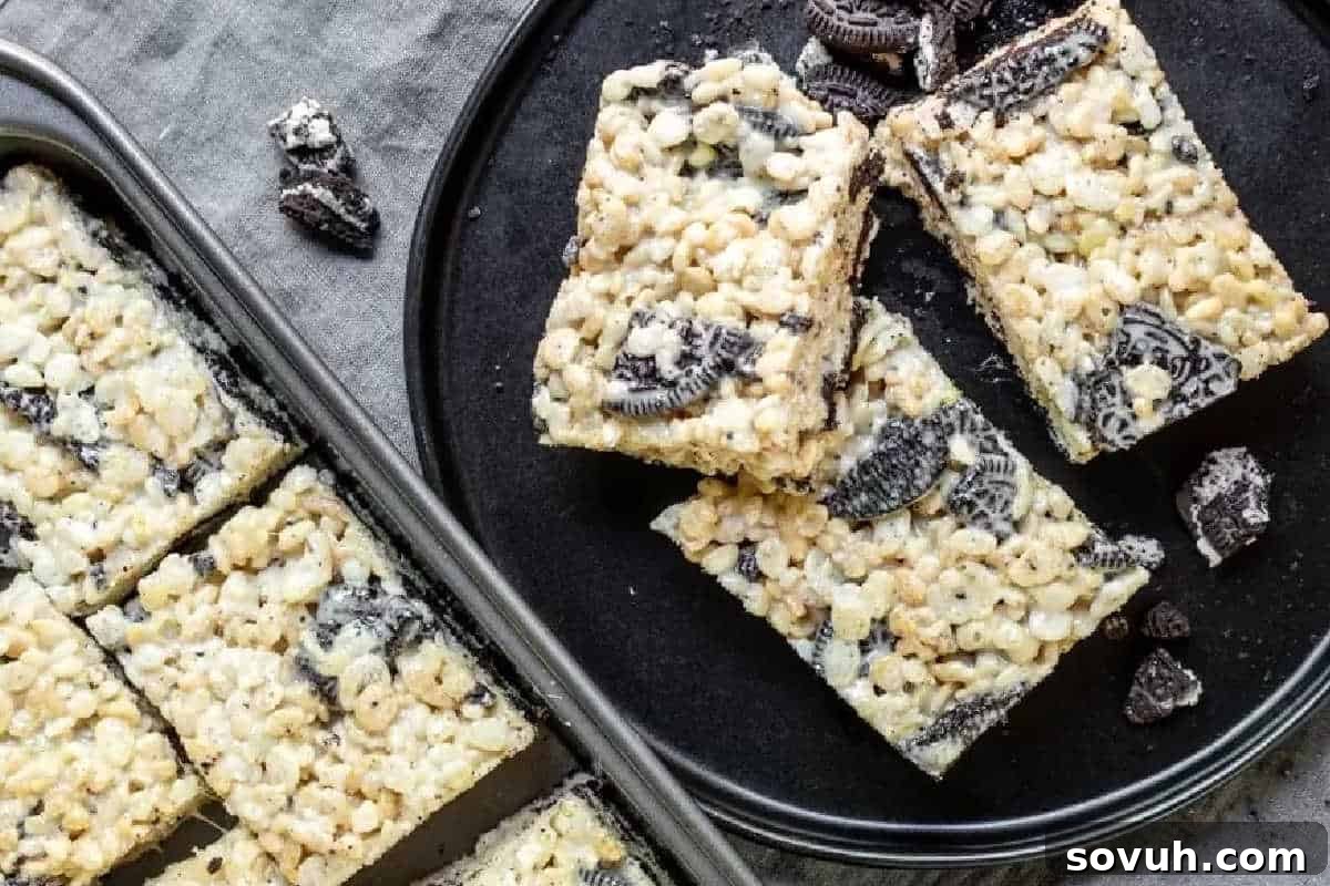 Delicious rectangular cookies and cream rice krispie treats, generously studded with crushed Oreo pieces, arranged artfully on both a black serving plate and within a practical baking tray.
