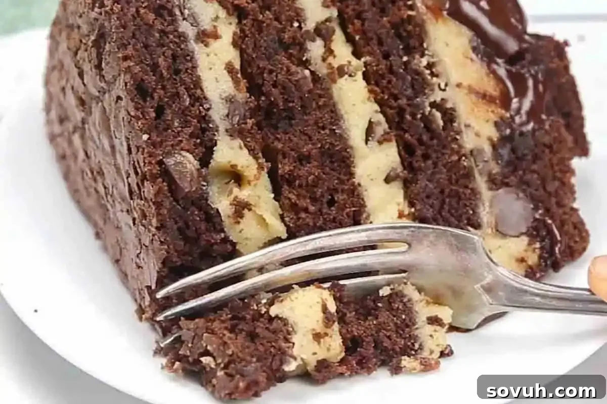 A fork is poised to cut into a luxurious slice of chocolate cake, revealing distinct layers of soft cookie dough and generous chocolate chips, elegantly presented on a pristine white plate.