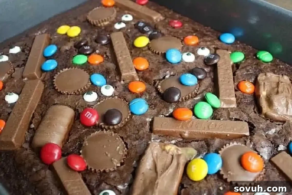 A full pan of rich brownies, artfully topped with an assortment of popular chocolate candies, including various peanut butter cups, classic chocolate bars, and vibrant candy-coated chocolates.