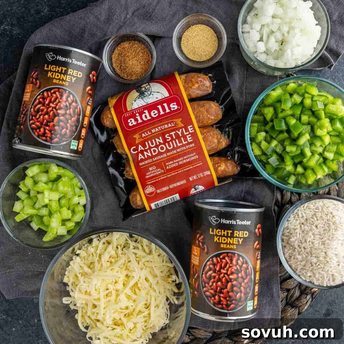 Top-down view of ingredients for a quick red beans and rice dish, featuring andouille sausage, red kidney beans, chopped onion, celery, green bell pepper, spices, shredded cheese, and rice.