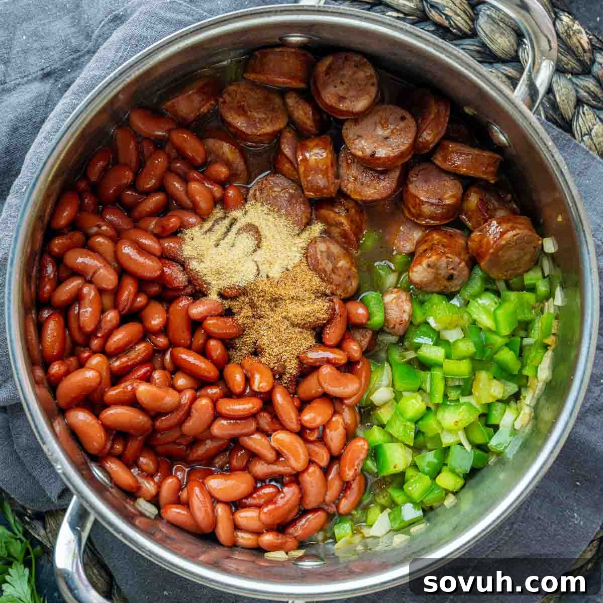 A pot containing sliced sausage, kidney beans, chopped green bell pepper, onions, and seasonings on a gray cloth—perfect for a quick red beans and rice dish.