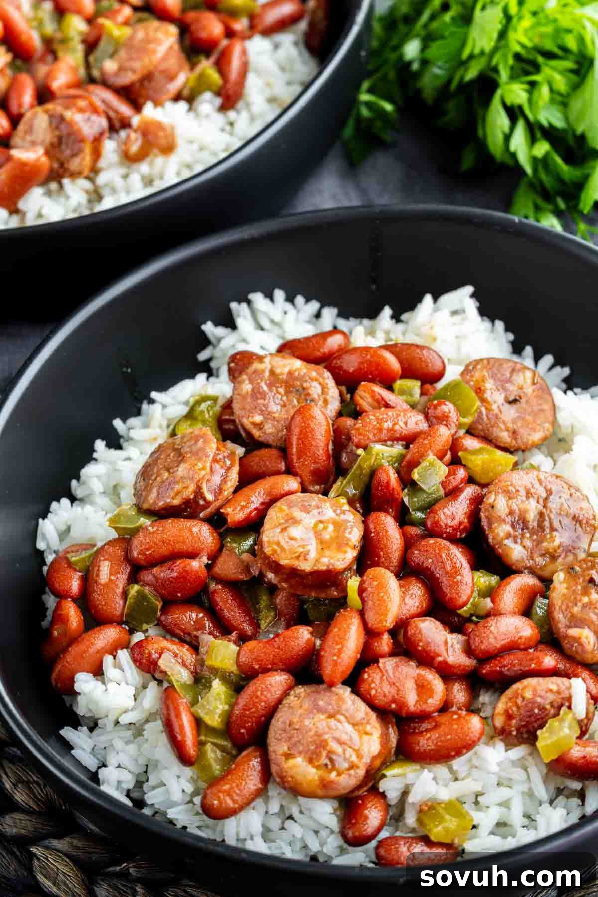 A bowl of white rice topped with red kidney beans, sliced sausage, and chopped green peppers—perfect for an easy red beans and rice recipe. Another bowl and fresh parsley are in the background.