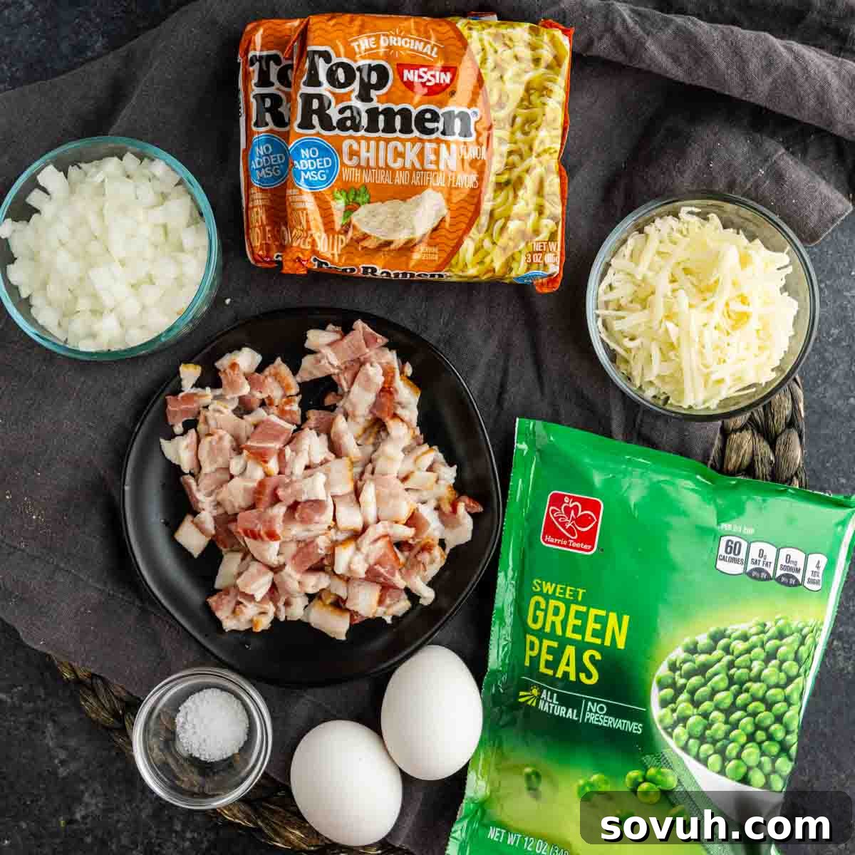 Effortless Ramen Carbonara 3 Ingredients including instant ramen noodles, chopped onions, shredded cheese, chopped bacon, frozen green peas, eggs, and a small bowl of salt arranged on a cloth for an Easy Ramen Carbonara.
