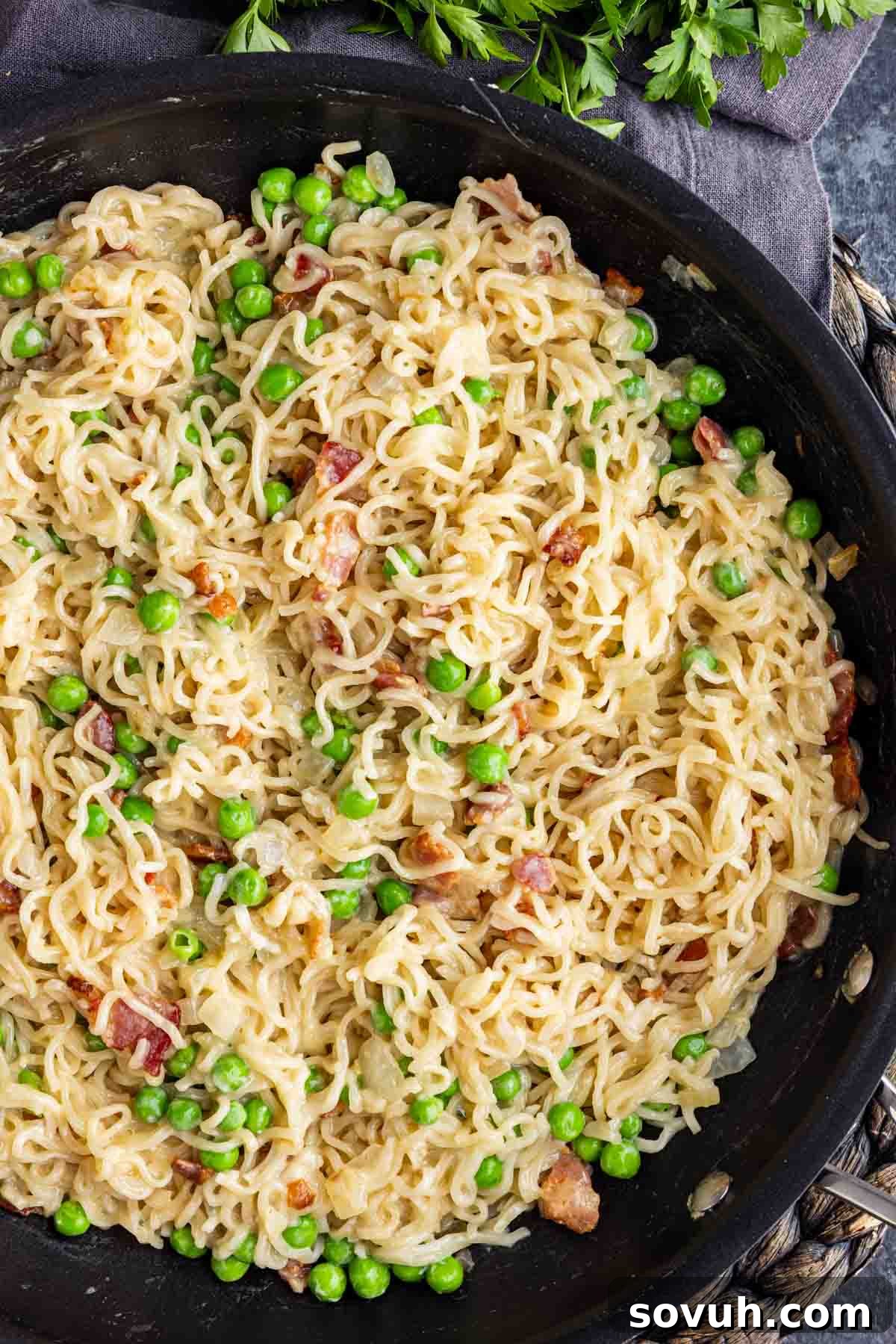 Effortless Ramen Carbonara 4 A skillet filled with creamy bacon ramen carbonara noodles mixed with green peas and pieces of bacon, garnished with fresh parsley.